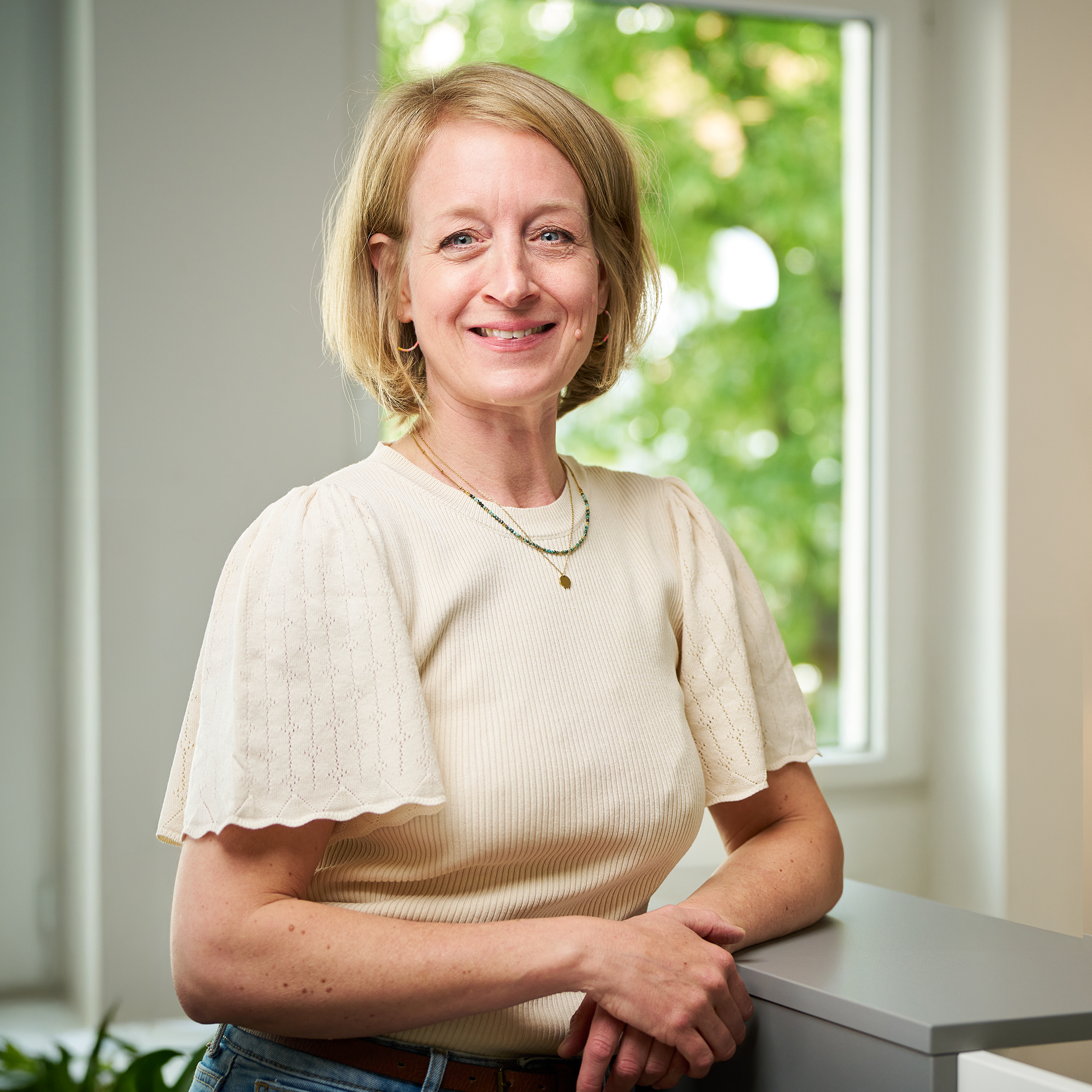 Eine lächelnde Frau steht entspannt an einem Tisch, trägt ein helles Oberteil, im Hintergrund Grünpflanzen.